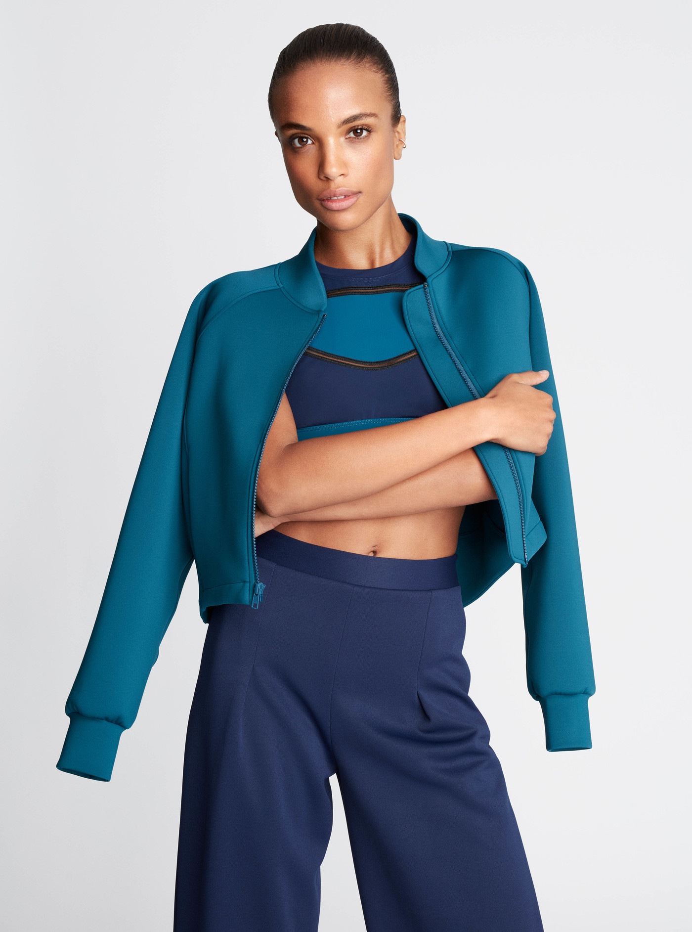 A brunette adult female poses in a teal jacket and navy blue pants against a white background in a fashion or beauty context.