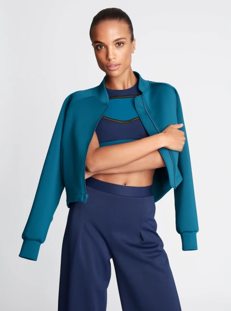 A brunette adult female poses in a teal jacket and navy blue pants against a white background in a fashion or beauty context.