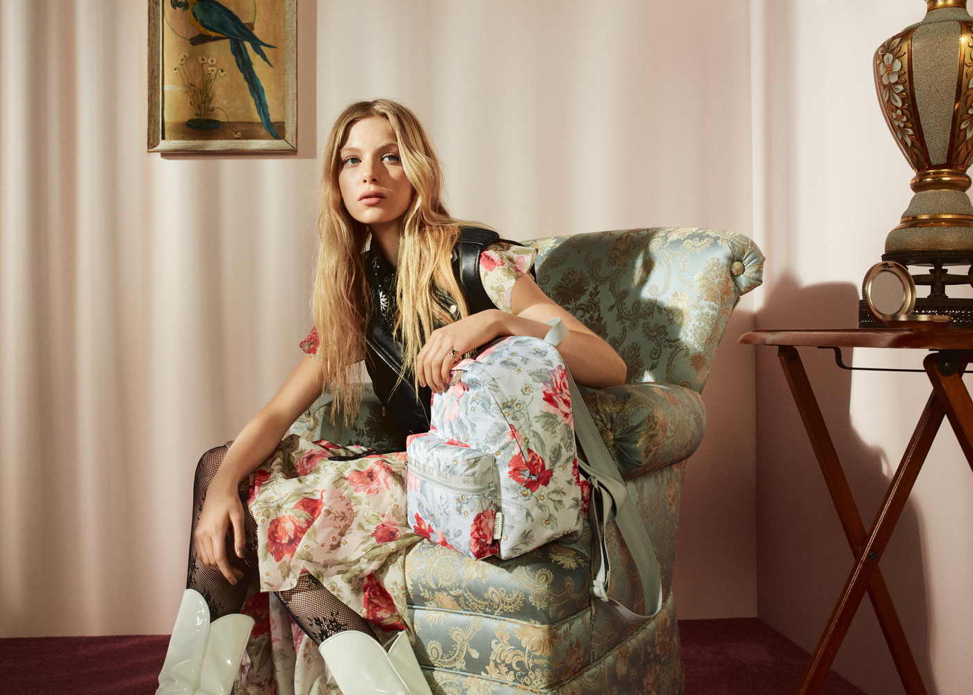 A blonde woman in a floral dress sits in a chair with a floral backpack, complemented by a vintage lamp, on a wooden table.