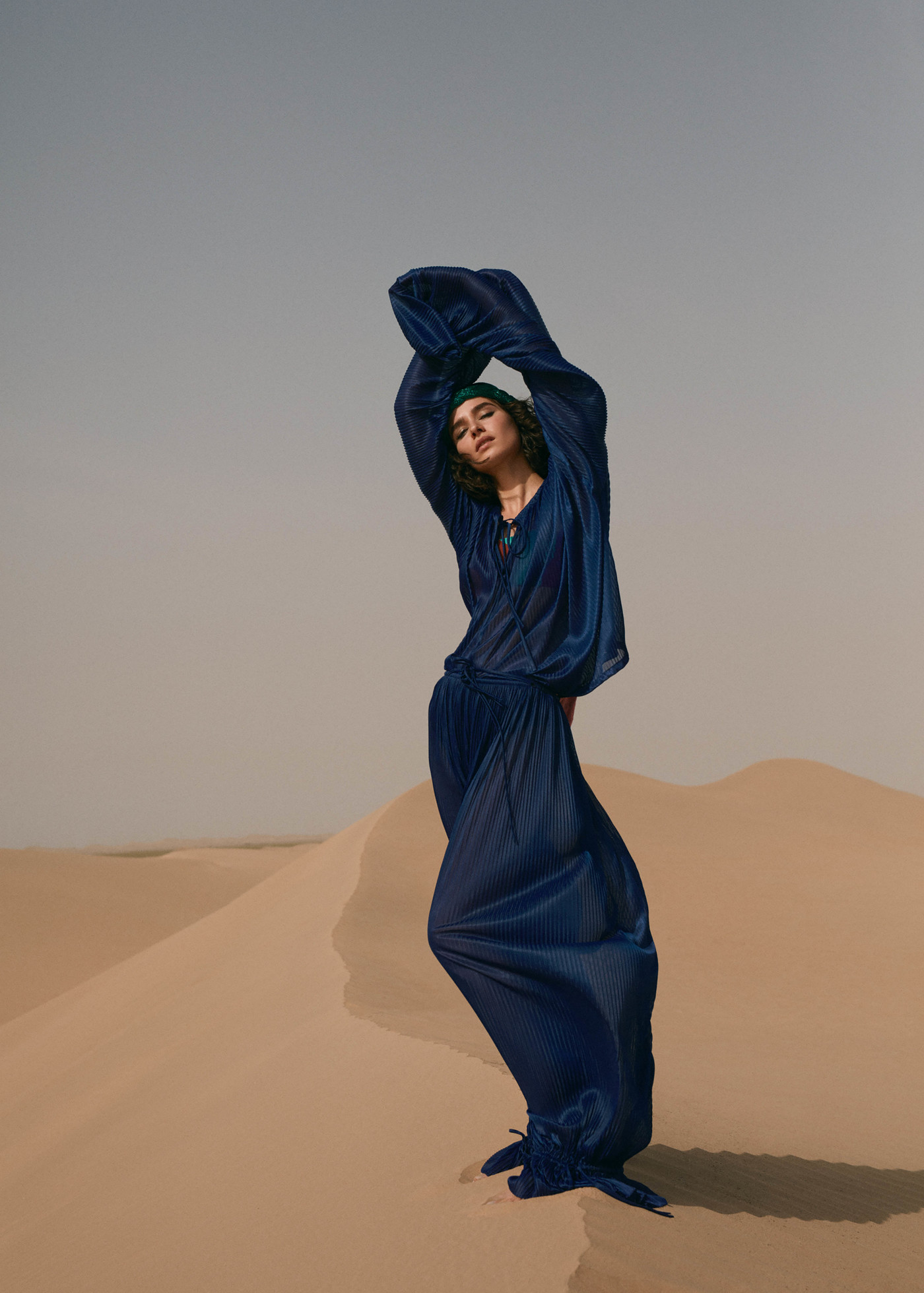 Fashionable woman in a blue pleated dress poses gracefully on a sand dune against a light blue sky, showcasing editorial style.