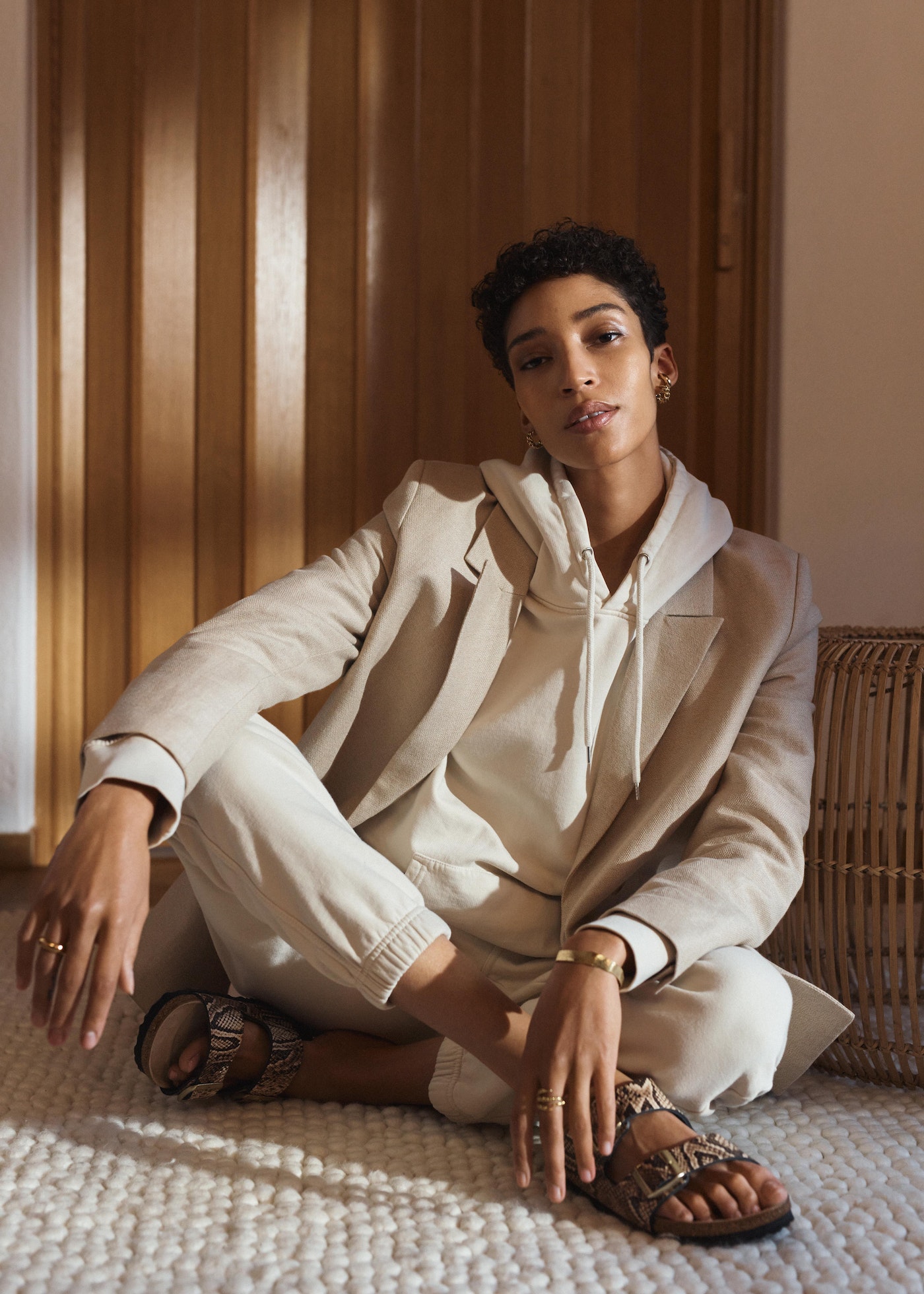 Young woman in a beige blazer and hoodie sits cross-legged on a textured rug. Neutral tones dominate the scene. Indoor setting.