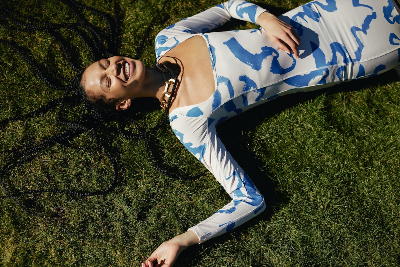 Woman in patterned dress smiles while lying on grass. Overhead shot captures long braids, necklace, and white-and-blue dress pattern.