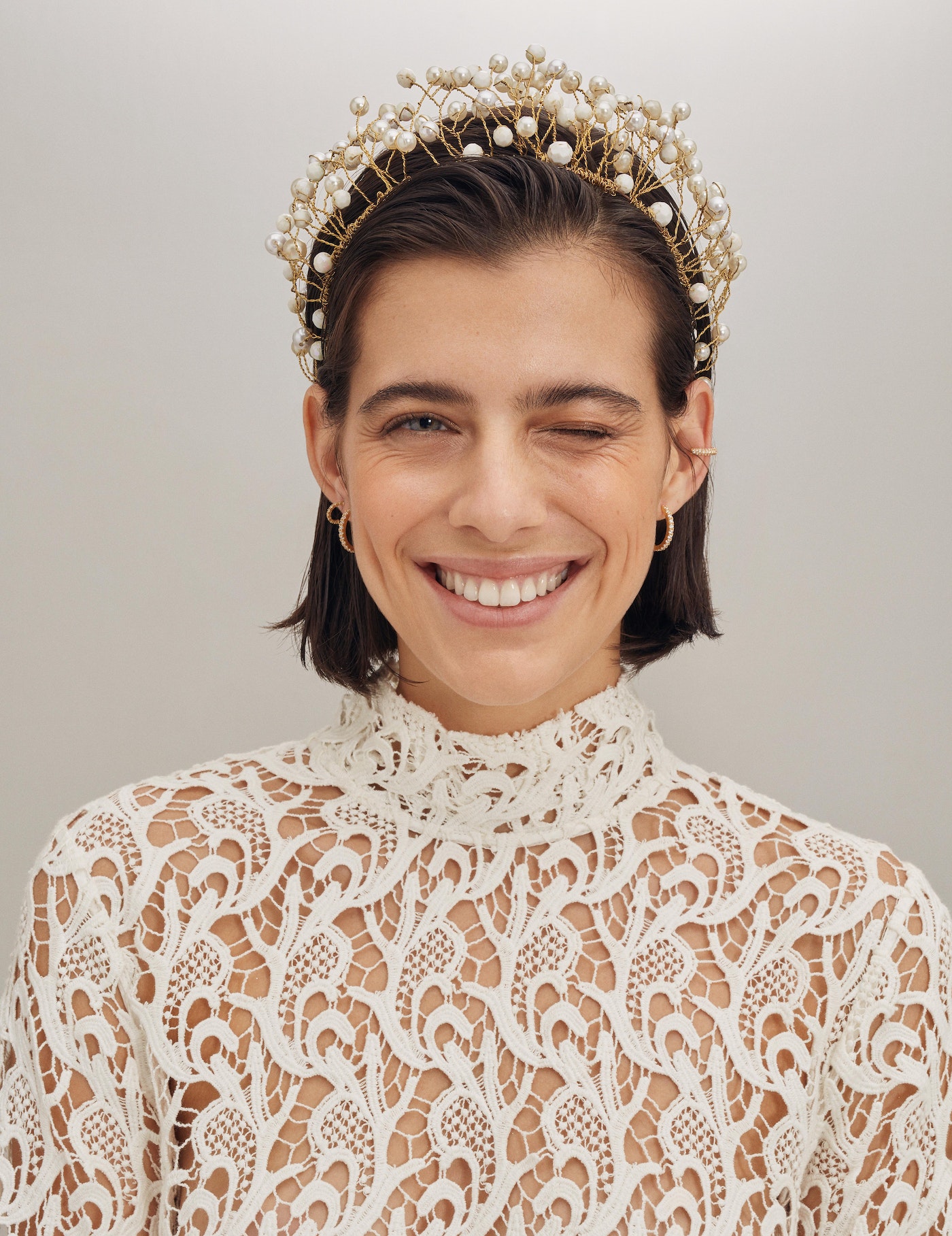 Close-up of a woman with short dark hair, fair skin, and a lace top, winking at the camera, wearing a decorative pearl headband.