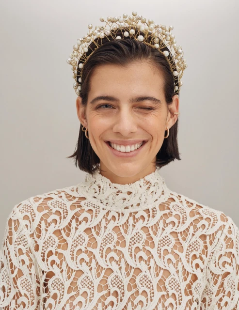 Close-up of a woman with short dark hair, fair skin, and a lace top, winking at the camera, wearing a decorative pearl headband.