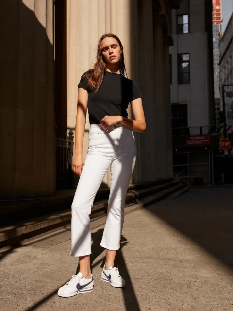 Woman in a black T-shirt and white jeans stands on an urban sidewalk in sunlight with buildings in the background. The shot is medium.