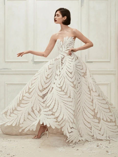 Woman in a white strapless gown posing in a neutral setting, showcasing high fashion and elegant design with soft, diffused lighting.