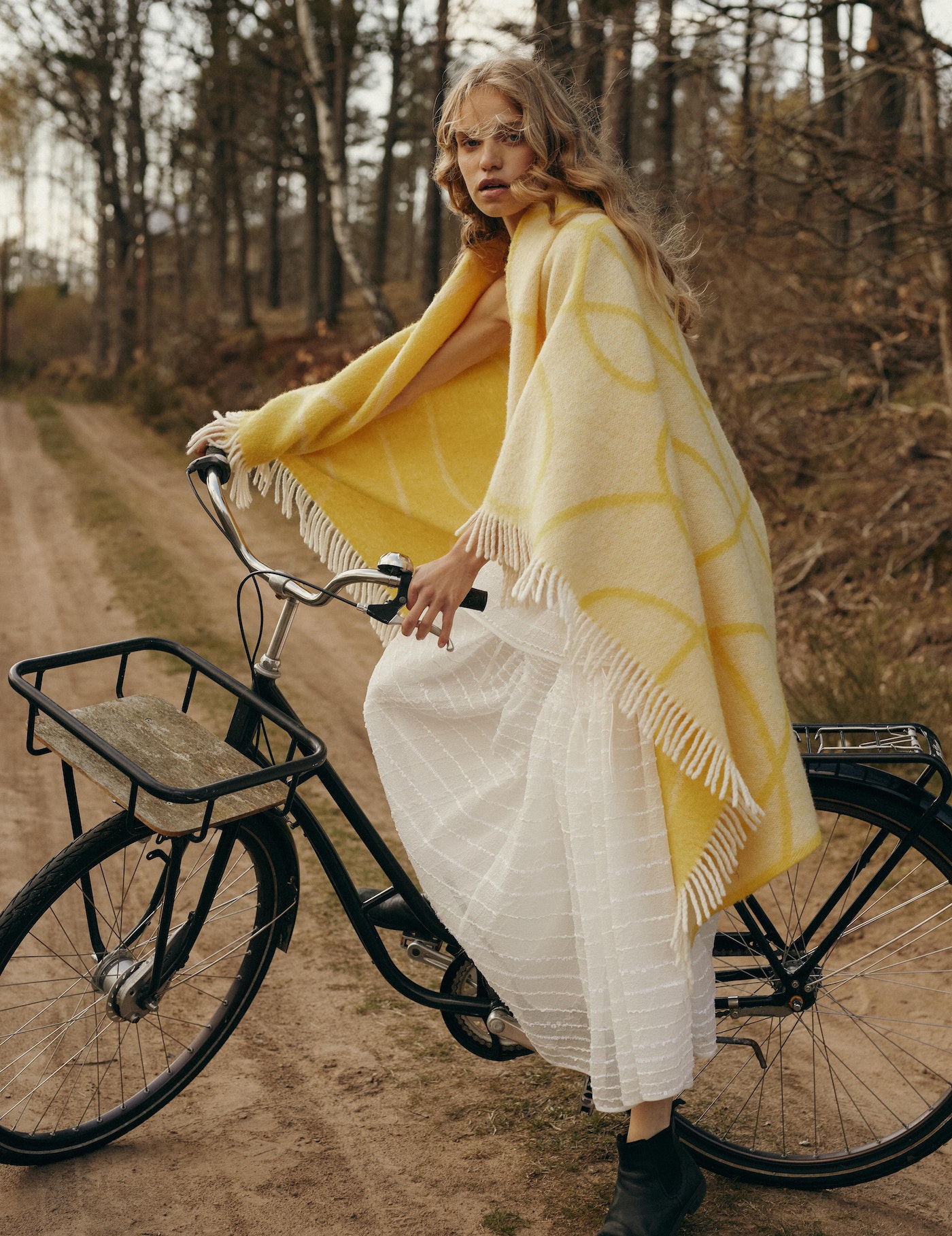 Woman in yellow blanket poses with a bicycle on dirt path in forest environment in lifestyle fashion shot with natural light.