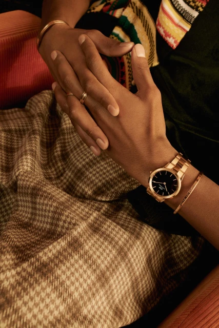 Close-up of two people holding hands, one wearing a rose gold watch and bangle bracelet, set against patterned textile backdrop.