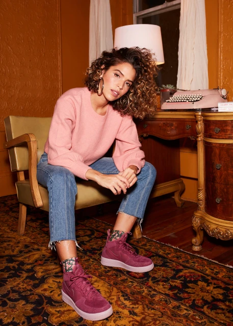Portrait of a woman with curly hair wearing a pink sweater and blue jeans sitting on a chair with a typewriter in the background.