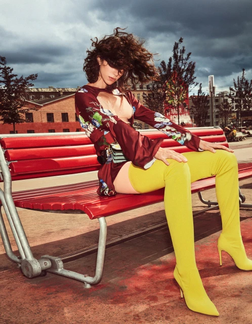 A woman with curly hair sits on a red bench wearing a floral top and yellow thigh-high boots against an urban outdoor backdrop with cloudy skies.