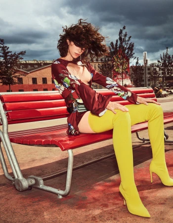 A woman with curly hair sits on a red bench wearing a floral top and yellow thigh-high boots against an urban outdoor backdrop with cloudy skies.