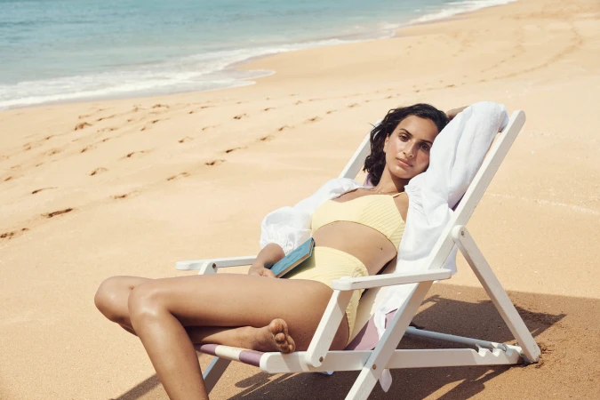 Woman in yellow bikini relaxing in a white beach chair on a sandy beach holding a book