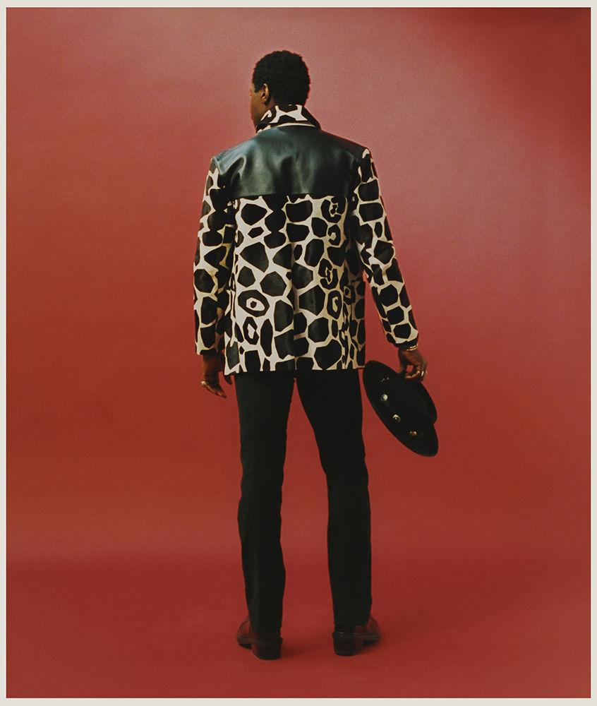 Man viewed from behind wearing a patterned animal print jacket and black trousers while holding a dark hat.
