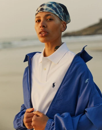 Model wearing a blue and white patterned headscarf, white polo shirt, and royal blue jacket outdoors near a beach.