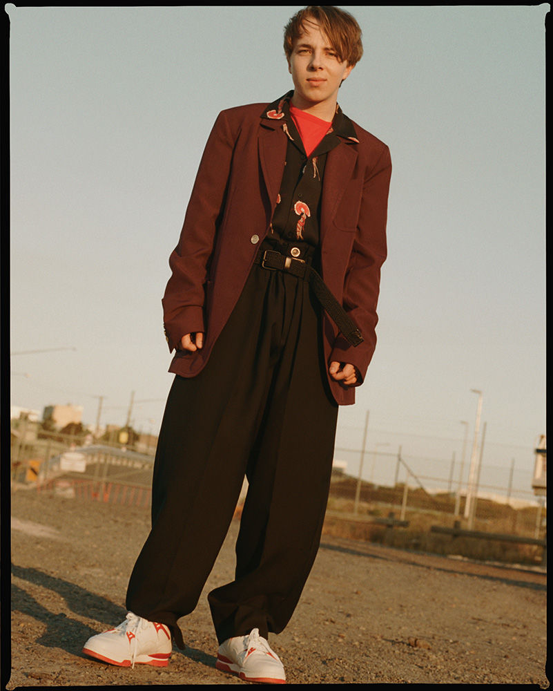 Stylish young adult posing outdoors in a maroon blazer, patterned shirt, black trousers, and white and red sneakers, set against a pale sky.