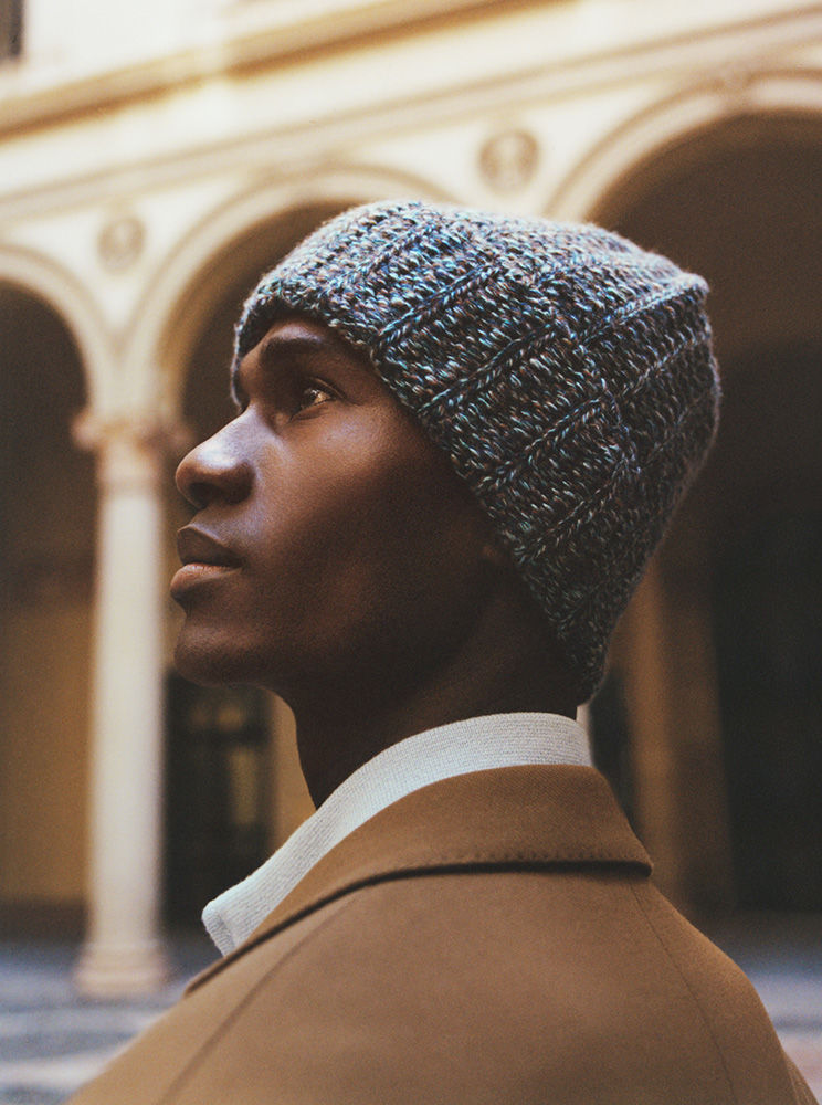 Portrait of a black man wearing a textured blue knit beanie and a camel coat against an arched courtyard background.