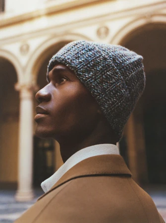 Portrait of a black man wearing a textured blue knit beanie and a camel coat against an arched courtyard background.