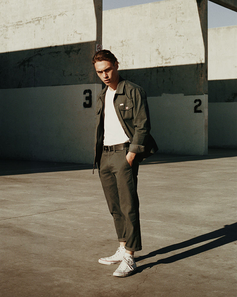 Young man in casual olive-green outfit stands on concrete; open jacket, white t-shirt, matching trousers, and white sneakers in sunlight.