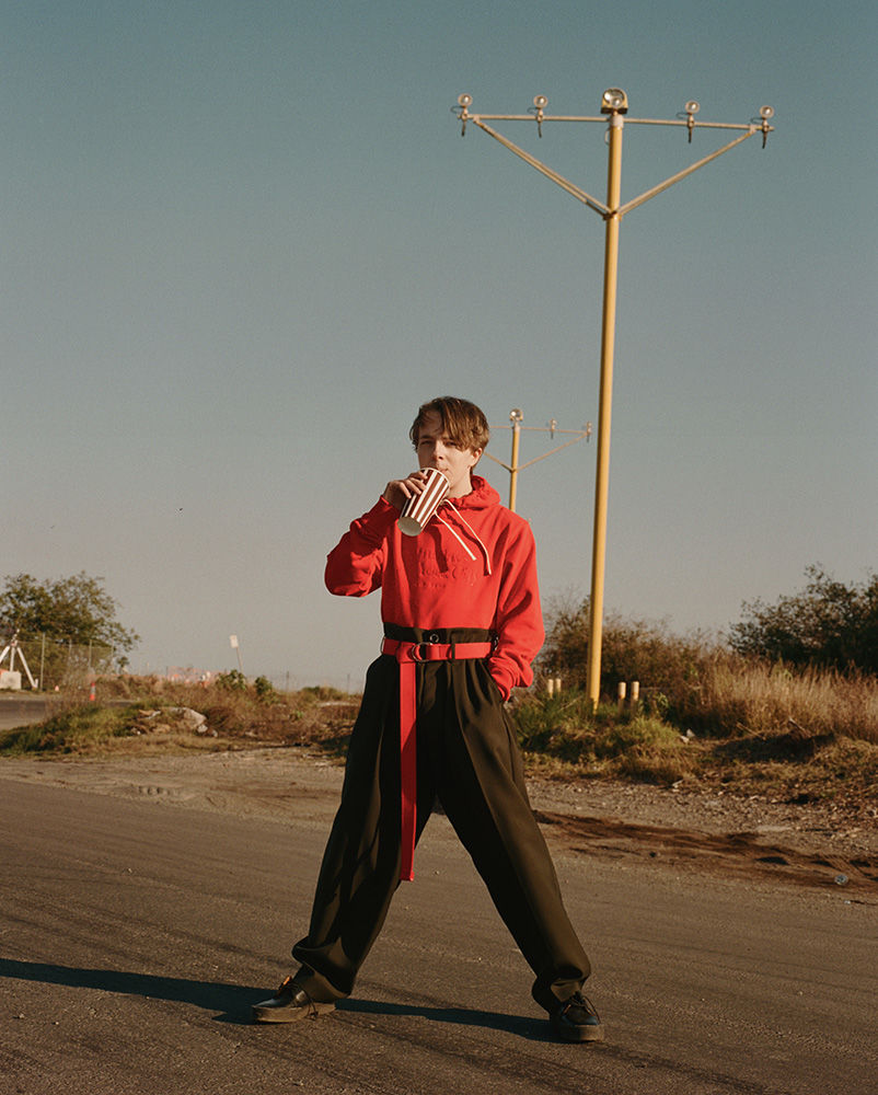 A person in a red hoodie and dark pants drinks from a striped cup on an empty road with utility poles under a clear blue sky.