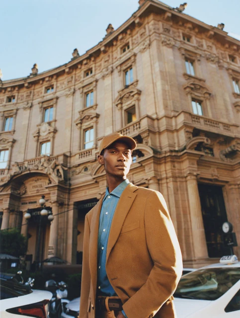 Man in a tan suit and baseball cap poses in an urban setting near a light-colored building and taxis, capturing a blend of fashion and city life.