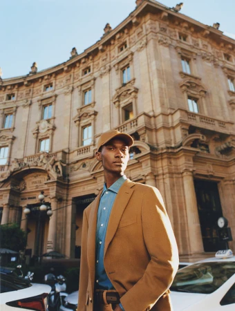 Man in a tan suit and baseball cap poses in an urban setting near a light-colored building and taxis, capturing a blend of fashion and city life.