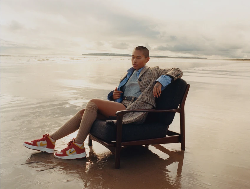 A woman in a blazer sits in a chair on a beach wearing sneakers in a fashion shot with waves and sky in the background.