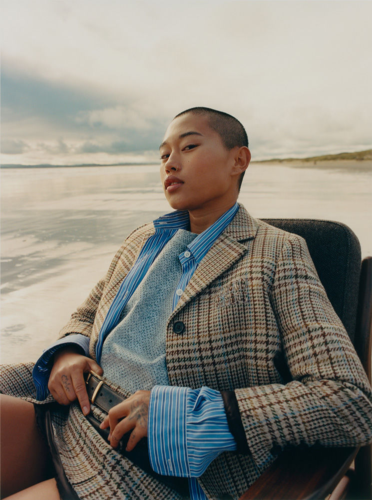 Woman in plaid blazer sitting on a chair on a beach. She is wearing a blue shirt and has short hair. Ocean and sky background.