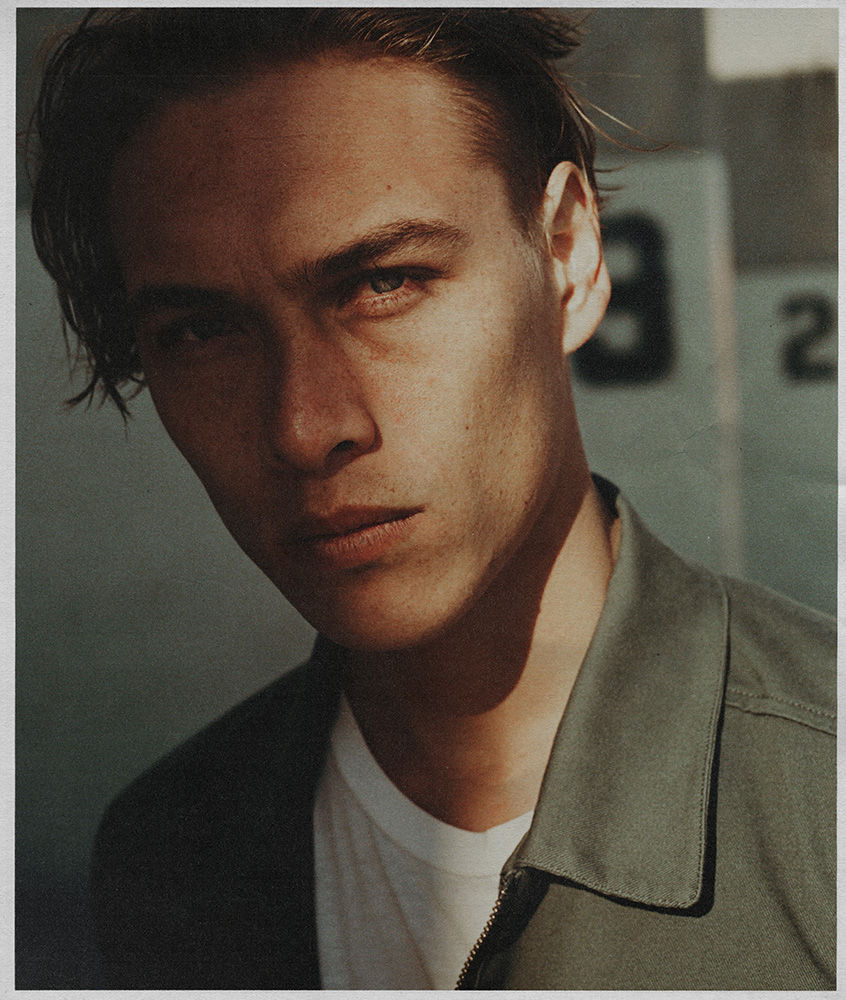 Portrait of a young man with brown hair and blue eyes, wearing an olive green shirt and white t-shirt. Light and shadows play across his face.