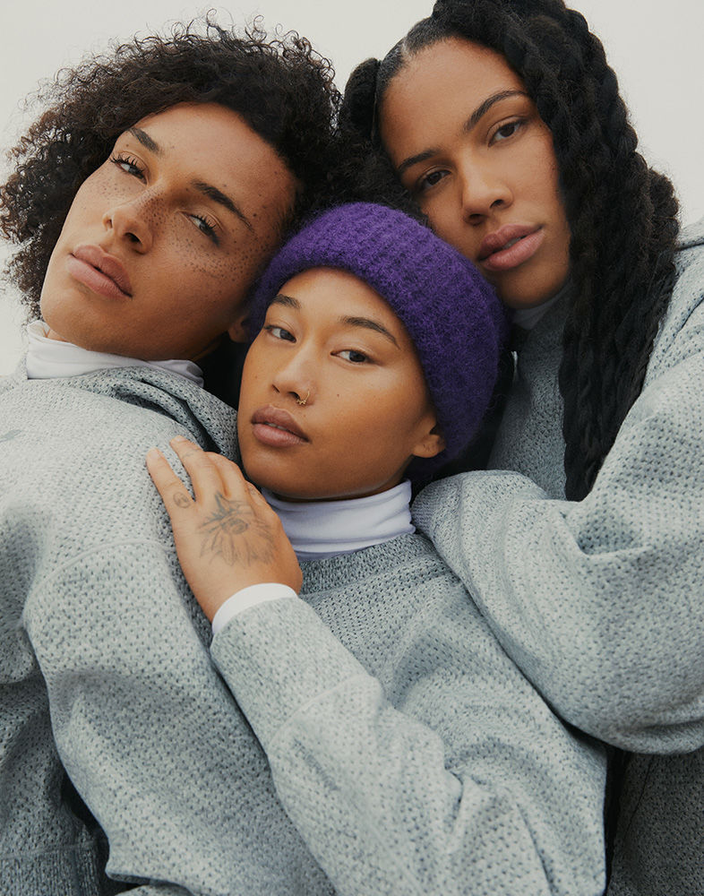 Three people with natural hair and grey sweatshirts pose in a studio. One wears a purple beanie in a portrait-style shot, indoor setting.