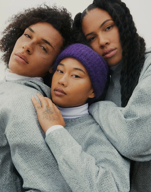 Three people with natural hair and grey sweatshirts pose in a studio. One wears a purple beanie in a portrait-style shot, indoor setting.