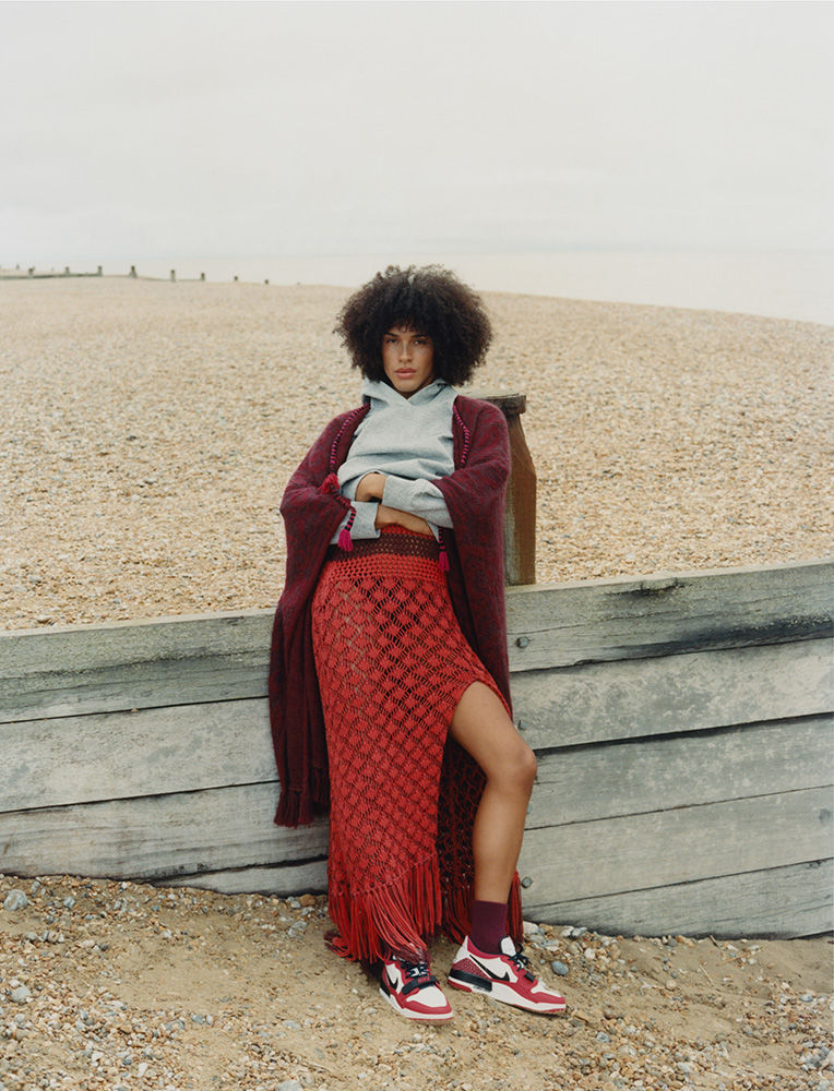 A stylish woman with an afro is posing outdoors wearing a grey hoodie, maroon cardigan, and a long, split red skirt on a beach.