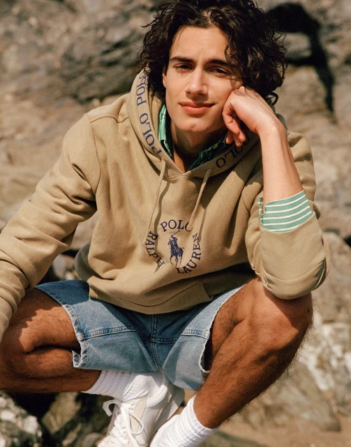 Young man in Polo Ralph Lauren hoodie and denim shorts sits casually outdoors on rocks, striking a relaxed pose with hand near face.