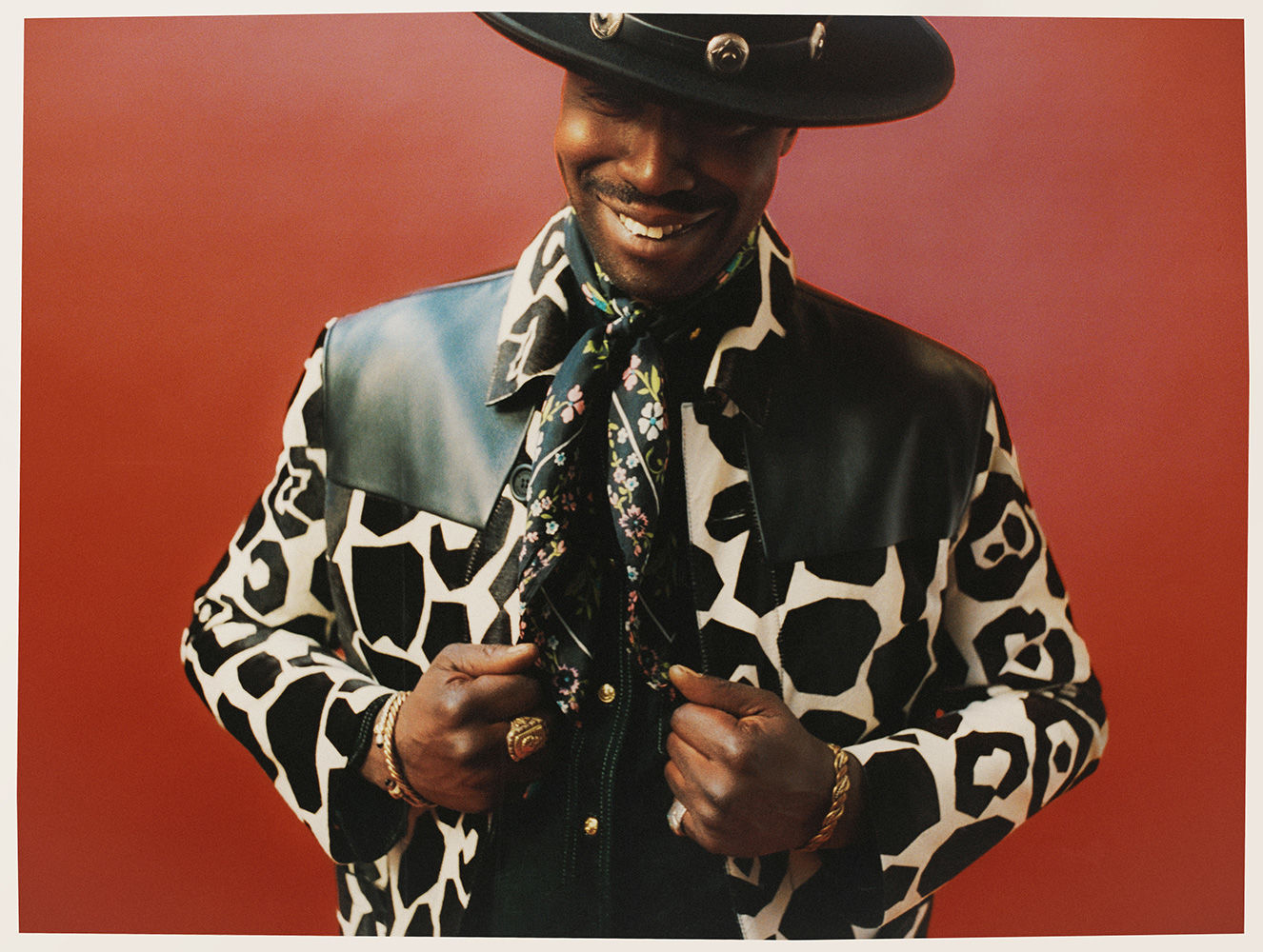 Stylish Black man in a cow-print jacket, black hat, and patterned scarf, smiling against a solid red background in a studio shot.