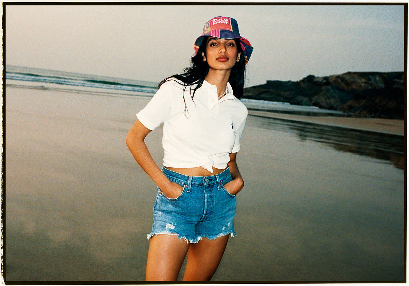 A woman in a bucket hat and denim shorts poses on a beach with dark sand and a misty sky. Fashion, summer, and beach lifestyle.