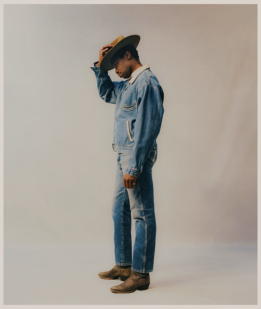 A stylish African-American man in a denim jacket and jeans poses in a photo studio, wearing a cowboy hat and boots against a grey background.