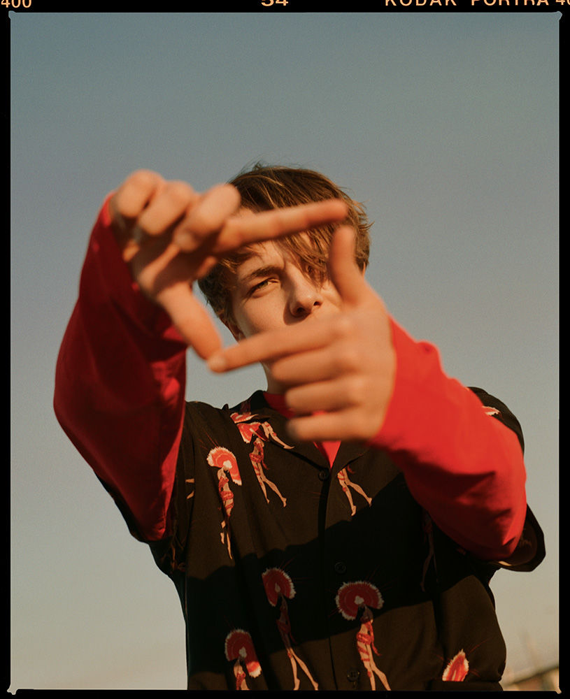 Portrait of a young man making a finger frame against a clear sky, wearing a patterned shirt and red undershirt, shot in natural light.