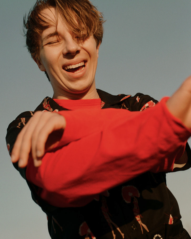 A young adult male with light brown hair and wearing a red long-sleeve shirt and patterned open button-down smiles against a clear blue sky.