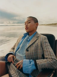 Close-up portrait of a person with a shaved head seated outdoors in a chair wearing a plaid blazer and blue striped shirt