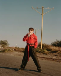 Young man in a red hoodie and black trousers drinking from a paper cup on an asphalt road with tall yellow light poles in the background.