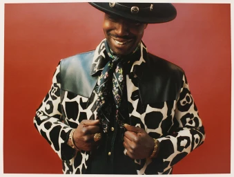 Black man wearing a black hat, patterned black and white jacket, and a floral scarf against a solid red backdrop