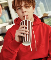 Young man with reddish-brown hair wearing a bright red hoodie while drinking from a striped paper cup with a straw
