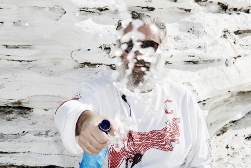 Man with beard in sunglasses sprays water from a blue bottle outdoors against a textured white wall, creating a splash.