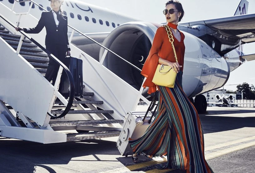 Two women on an aircraft stairs: one is a flight attendant in uniform carrying a suitcase, and the other is a fashion model in stylish outfit.