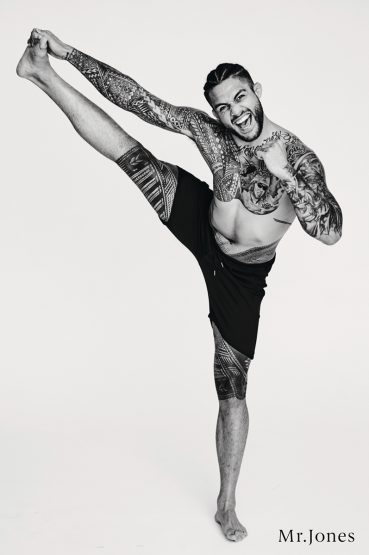 Athletic man with tribal tattoos performing a high kick against a white background. He is smiling, shirtless, and wearing dark shorts.