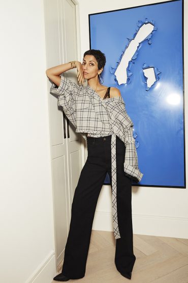 A woman in a plaid shirt and black pants poses indoors against a blue abstract artwork, illuminated by bright lighting.