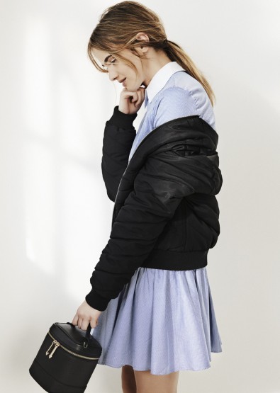 An adult woman in a light blue dress and black jacket stands against a white background, holding a black round vanity case.