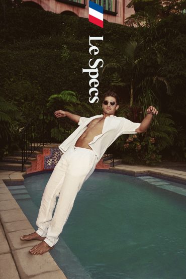 Young adult male poses at the edge of a pool in a white outfit, next to greenery and a partial building, summer mood, daytime.