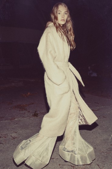 A fair-skinned woman with long blonde hair wears a light beige belted coat and flared pants while walking on a cement surface at dusk.