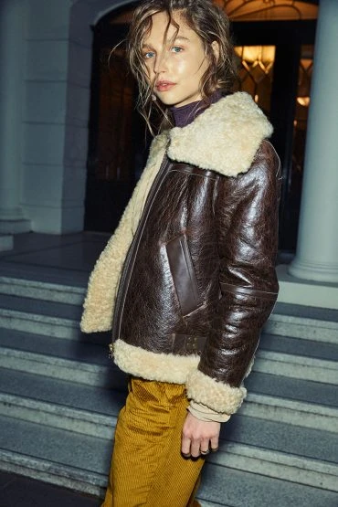 Blonde woman in a brown leather aviator jacket with a cream-colored fur collar and yellow corduroy pants poses on concrete stairs.