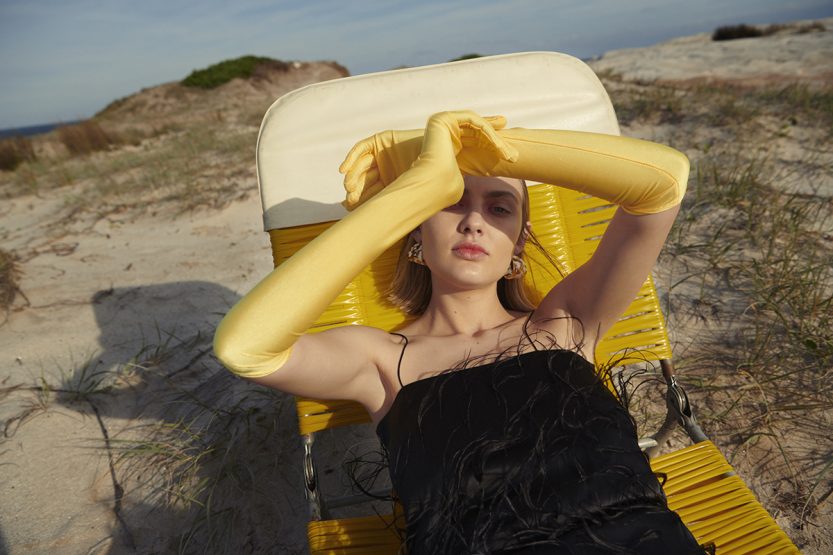 Young woman in a black dress and yellow gloves reclines on a yellow lounger on a beach with sand dunes under a bright sky.